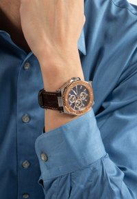 Reloj con correa de cuero marrón, con caja octagonal en oro rosa, esfera negra y acentos plateados, llevado en una muñeca debajo de una camisa azul.