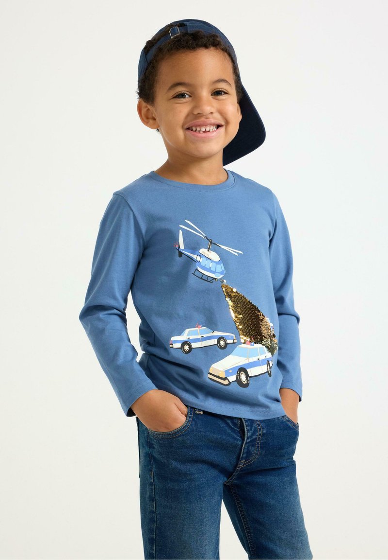 Blå langærmet skjorte med grafik af en helikopter og en polisecar, med detaljer i guldpalietter, parret med blå jeans og en mørk kasket.