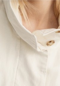 Giacca color crema con colletto, dal tessuto morbido, caratterizzata da una chiusura con bottone prominente e un tessuto liscio, che enfatizza il design minimalista.