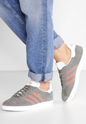 Osoba w podwiniętych niebieskich dżinsach i szarych zamszowych trampkach z różowymi paskami i białymi podeszwami, idąca po białej powierzchni.