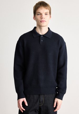 Pull en tricot côtelé bleu marine avec un col, trois boutons et des manches longues. Le tissu a une apparence texturée et une bordure ajustée.