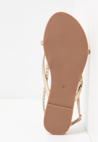 Płaskie sandały z jasnobrązową podeszwą, złotymi plecionymi paskami oraz rozmiarem 37 wytłoczonym na podeszwie, umieszczone na białym tle.