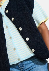 Gilet in felpa navy con superficie testurizzata, dotato di bottoni color crema, indossato sopra un top in maglia a righe blu chiaro e giallo.