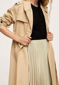 Trench-coat beige avec une taille à ceinture, des manches retroussées et des épaulettes aux épaules, superposé sur un haut noir à côtes et une jupe plissée vert menthe.