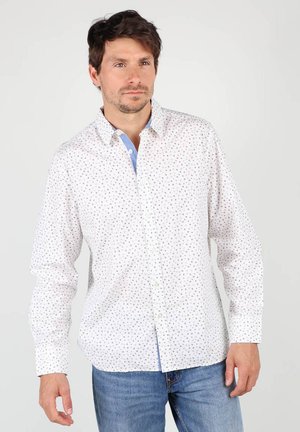 Chemise blanche à manches longues avec un motif de points bleus et verts, un col intérieur bleu contrasté, accompagnée d'un jean en denim bleu.