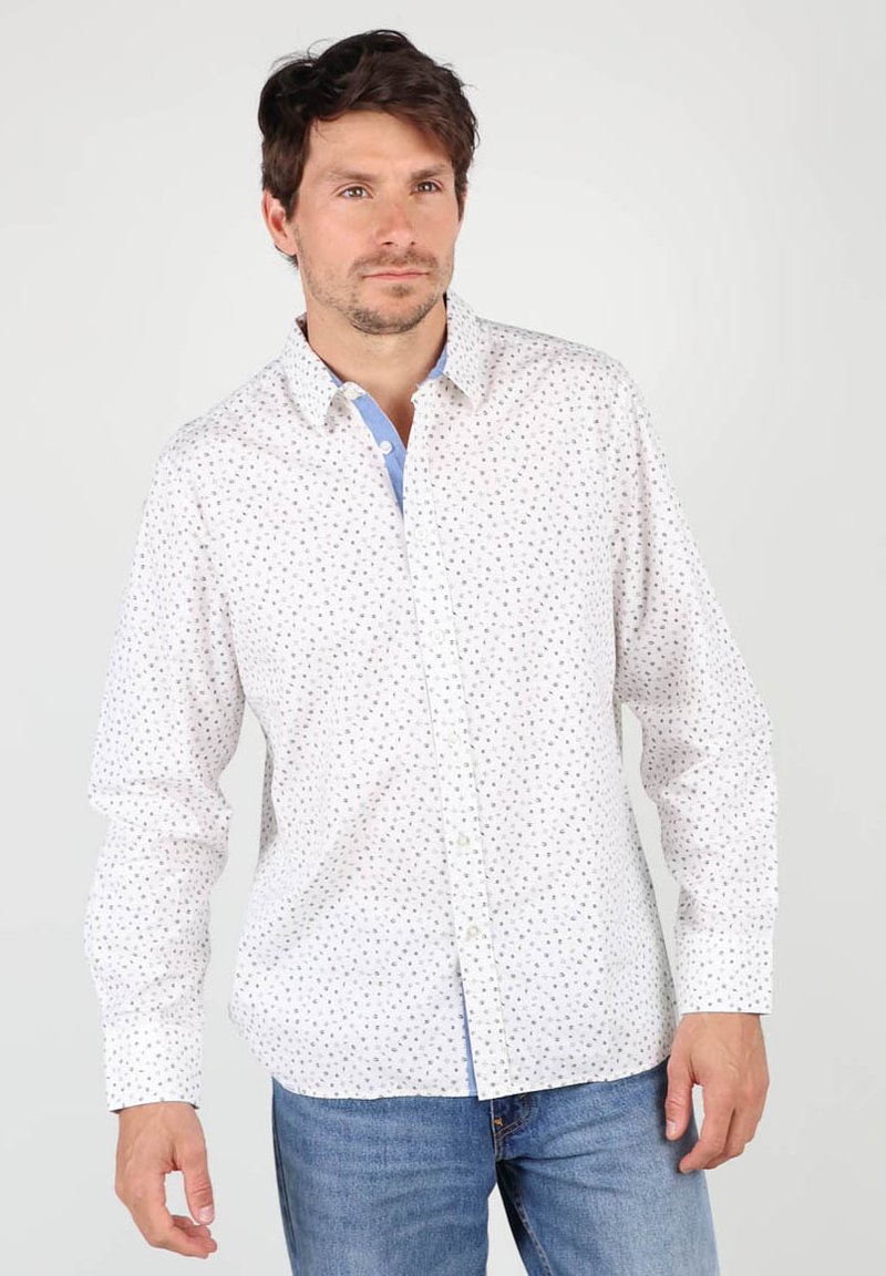 Chemise blanche à manches longues avec un motif de points bleus et verts, un col intérieur bleu contrasté, accompagnée d'un jean en denim bleu.