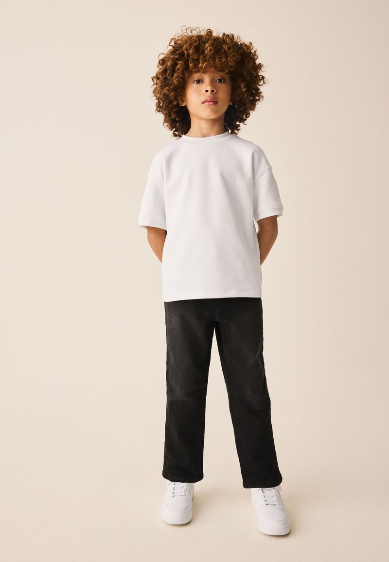 Barn med krøllet hår står med fronten mod kameraet iført en ensfarvet hvid t-shirt, sorte bukser og hvide sneakers mod en beige baggrund.
