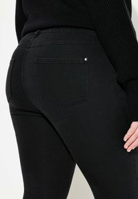 Jeans de denim negro con un ajuste slim, dos bolsillos traseros y un pequeño acento metálico en el bolsillo derecho. Textura suave, diseño de cintura media.
