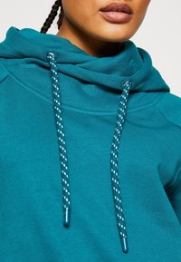 Sweat-shirt à capuche bleu sarcelle avec un col bénitier, des cordons de serrage à pois blancs, et un tissu doux et texturé avec des détails de couture visibles.