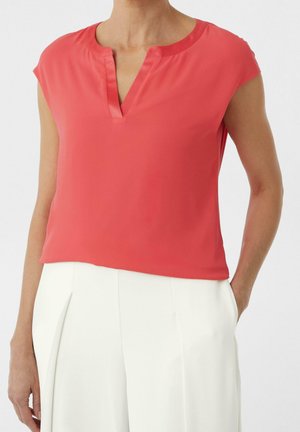 Kvinde iført en ærmeløs koralfarvet V-hals bluse kombineret med højtaljede hvide bukser, stående med den ene hånd i lommen.