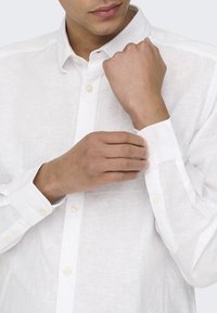 Camisa blanca de lino con cuello abotonado y mangas largas. Presenta una textura sutil y un cierre clásico de botones en la parte delantera.