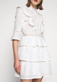 Valkoinen mekko, jossa on lyhyet hihat, röyhelökoristeet ja kerrokselliset osat. Valmistettu kirjaillusta kankaasta, jossa on kukkakuosisia ja leikattuja yksityiskohtia.