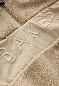 Bežinės tekstūruotos fliso striukės su lygiomis detalėmis; turi išsiuvinėtą "BAKER" logotipą ir prekės ženklą "by TED BAKER."