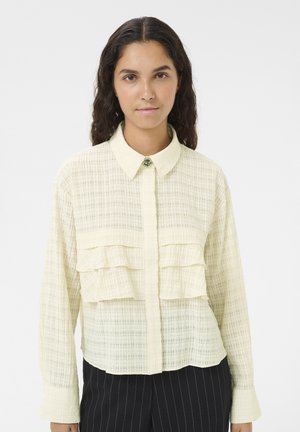 Blusa gialla chiara con tessuto texture, dotata di una chiusura frontale con bottoni, colletto e due volants sovrapposti sul petto. Maniche lunghe.