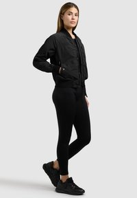 Vrouw staand met één hand in de zak, gekleed in een zwart bomberjack, zwarte legging en zwarte sneakers tegen een effen lichte achtergrond.