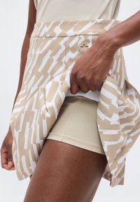 Χέρι σηκώνει μπεζ και άσπρη φούστα με σχέδιο, αποκαλύπτοντας μπεζ εφαρμοστά σορτς από κάτω, σε ουδέτερο φόντο.