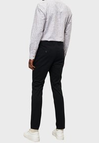 Pantaloni neri con una vestibilità su misura, dotati di due tasche posteriori, abbinati a una camicia a maniche lunghe con un motivo. Indossati con sneakers bianche.