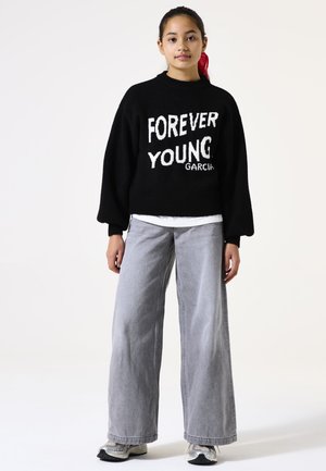 Pull en tricot noir avec "FOREVER YOUNG" en blanc, manches larges, associé à un jean léger gris taille haute et des baskets sportives.