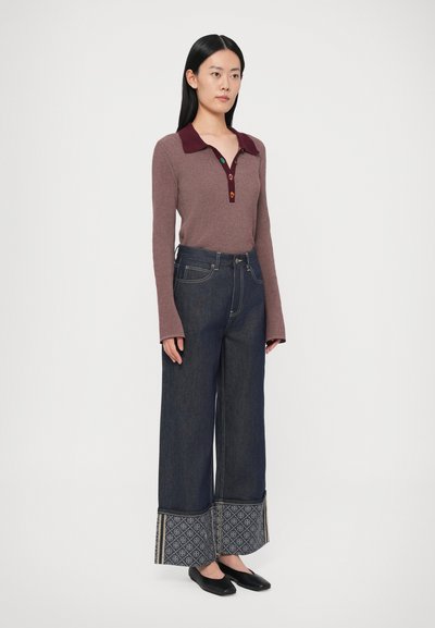 Garām piedurknēm adīts tops purpursārtā krāsā ar burgundijas krāsas apkakli un pogu plāksteri, kopā ar platām džinsa biksēm ar rakstainiem manšetiem.