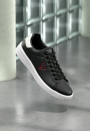 Schwarzer Sneaker mit weißer Sohle und Fersenlasche, rotes dreieckiges Logo an der Seite, schwarze Schnürsenkel, vor einer Glassteinwand dargestellt.