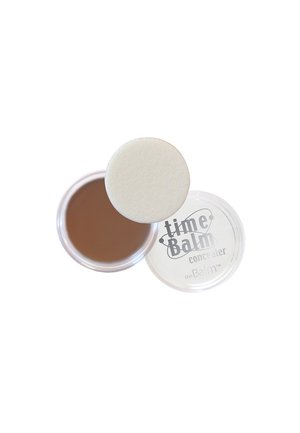 Brun krämig concealer i en genomskinlig rund behållare med en vit puffapplikator. Locket har en etikett med texten "timeBalm concealer" i grått.
