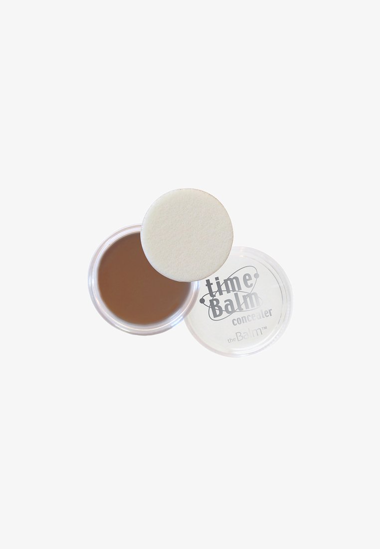 Brun krämig concealer i en genomskinlig rund behållare med en vit puffapplikator. Locket har en etikett med texten "timeBalm concealer" i grått.