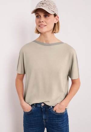 Kvinde iført beige kortærmet top, blå jeans og beige mønstret kasket, stående med hænderne i lommerne mod en ensfarvet baggrund.