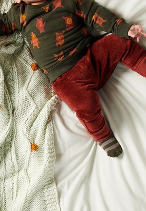 Tout-petit allongé sur le lit portant un pull vert avec des ours orange, un pantalon rouge, des chaussettes rayées, à côté d'une couverture en tricot crème avec des pompons orange.