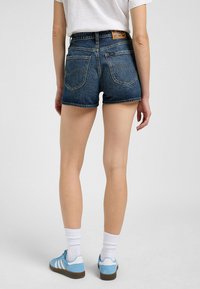 Shorts de denim de talle alto en azul oscuro con un dobladillo deshilachado, que cuentan con dos bolsillos traseros y una etiqueta de cuero marrón en la cinturilla.