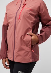 Giacca impermeabile rosa con tasca con zip e cappuccio regolabile. Presenta maniche scure a contrasto e polsini elastici. Materiale leggero.