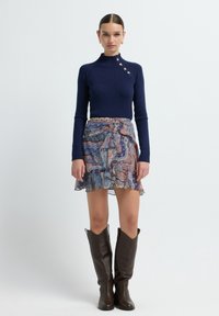 Marineblauwe geribbelde trui met knoopdetails, gecombineerd met een met patroon versierde flared rok in blauwe en bruine tinten. Bruine kniehoge laarzen maken de outfit compleet.