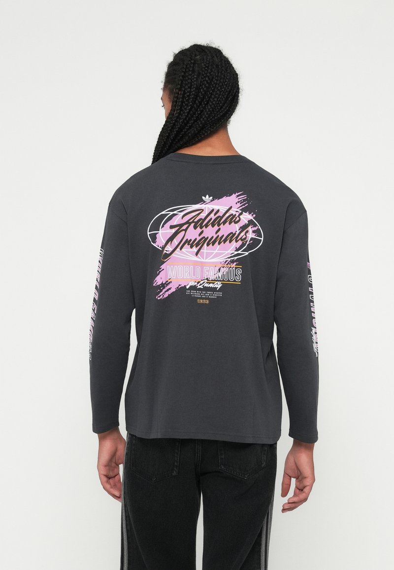 Person mit langen Zöpfen, der ein schwarzes Adidas Originals Langarmshirt mit pinken und weißen grafischen Texten auf dem Rücken und den Ärmeln trägt.