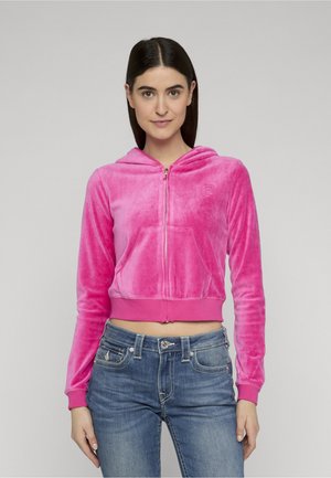 Donna che indossa una felpa con cappuccio in velluto rosa acceso con cerniera e jeans blu, in piedi contro uno sfondo chiaro e uniforme.