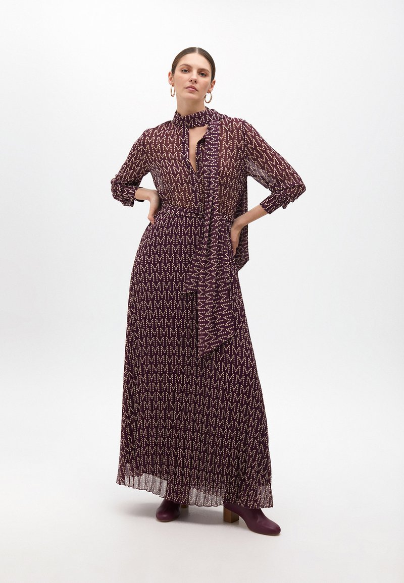 Vestido maxi de tela morada oscura con un patrón en M beige, con mangas largas, cuello con lazo y una silueta fluida. Combinado con botas de tobillo moradas.