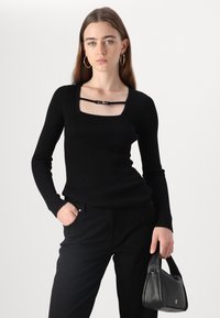 Zwarte geribbelde longsleeve top met vierkante halslijn en zwarte hardware details. Gepaard met zwarte broek en een kleine zwarte handtas.