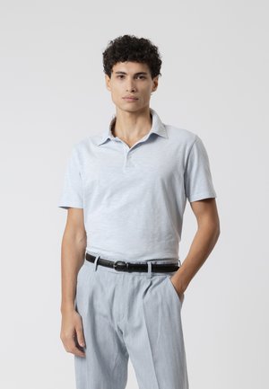Joven con cabello rizado que lleva una camisa polo azul claro metida en unos pantalones a juego con cinturón negro, de pie con una mano en el bolsillo.