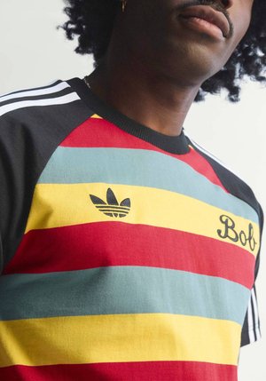 Persoană purtând un tricou în dungi multicolore cu mâneci negre, logo-ul Adidas și textul „Bola” pe piept, văzută de sub bărbie.