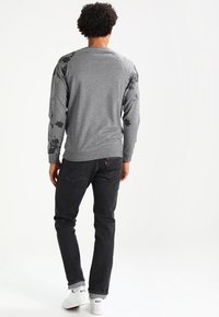 Sudadera gris con patrón floral en las mangas, puños y dobladillo acanalados; combinada con jeans negros y zapatillas blancas.