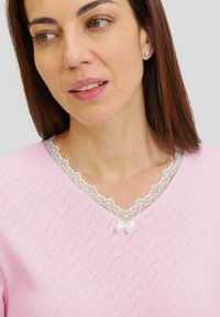 Top de color rosa claro con escote en V que presenta un patrón de chevron texturizado, un cuello de encaje blanco y un pequeño lazo blanco como acento en el escote.