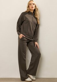 Vrouw met lang haar, gekleed in een bruine ritsvest en bijpassende wijde broeken, staande tegen een beige muur met één hand in de zak.