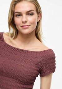 Haut maroon à épaules dénudées avec un tissu texturé et froncé, manches courtes et coupe ajustée. Peau lisse et maquillage naturel avec des cheveux blonds.