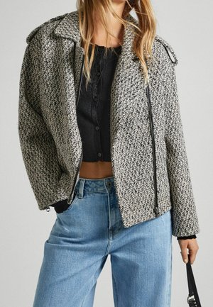Kvinde iført sort kropsnær knap-top, grå struktureret lynlåsjakke og lyseblå højttaljede jeans, der holder en sort skuldertaske.