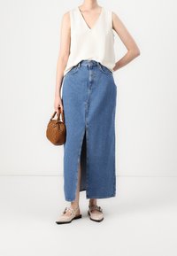 Denim maxi skirt with a front slit, high waist, and blue color. Paired with a beige sleeveless top and light-colored flat shoes. Accessories include a small brown handbag.