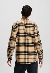 Camisa de cuadros beige de franela, con un patrón de rejilla negra, mangas largas y puños de botón. Etiqueta en la parte inferior izquierda.