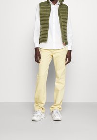 Grön puffy väst, vit skjorta med knappar, gula byxor och grå-vita sneakers. Slät textur, lätta material och avslappnad passform.