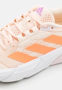 Chaussure de sport pêche clair avec dessus en mesh, bandes orange contrastées, semelle blanche et logo rose sur la languette et le côté.