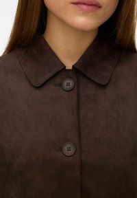 Žena nosi tamnosmeđi suđeni kaput s šiljastim kragnom i dva velika crna dugmeta zakopčana sprijeda.