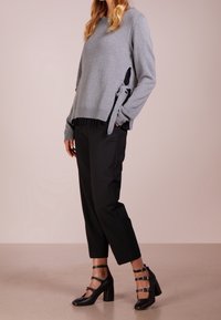 Grå stickad tröja med sidoknyten, svart plisserad underlayer synlig vid fållen, tillsammans med svarta skräddarsydda byxor och svarta sandaler med remmar.