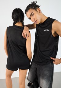 Schwarze, durchbrochene Tanktops mit abgerundetem Saum, die auf der Vorderseite den Text "NEW YORK" tragen; kombiniert mit zerstörten Denim-Shorts und Jeans.