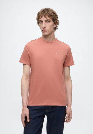 Νέος άνδρας με σγουρά μαλλιά, ντυμένος με ένα απλό ροζ σολομού κοντομάνικο μπλουζάκι και σκούρα μπλε τζιν, στέκεται μπροστά σε ένα φωτεινό φόντο.